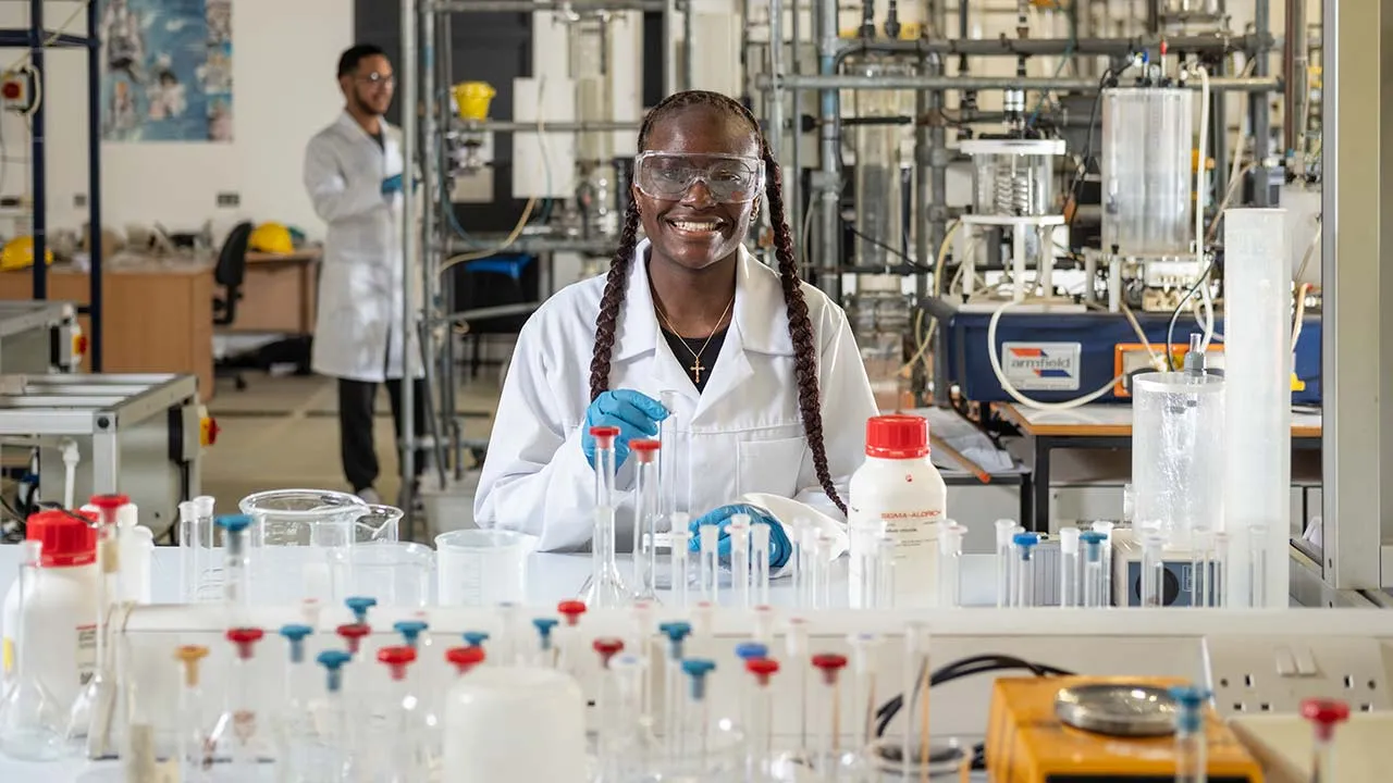 A student wearing a lab coat and safety goggles is working on a chemical experiment. There are lots of glass beakers and test tubes on the bench in front of them