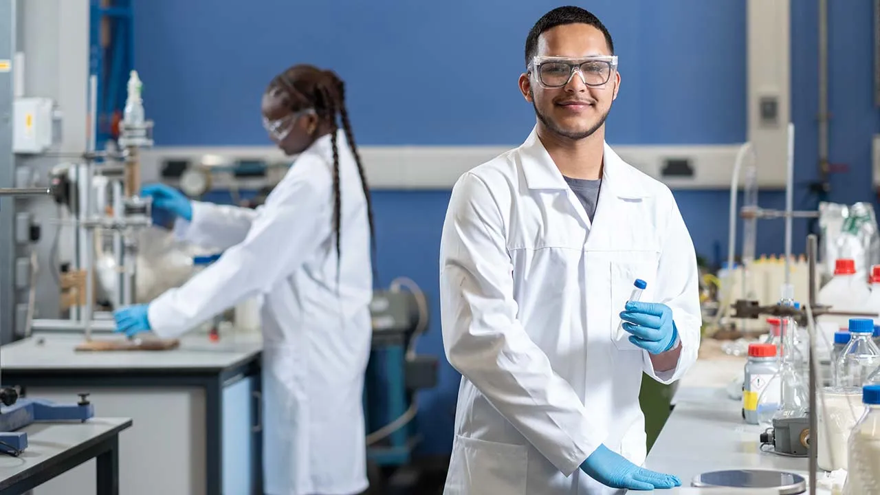 Two people in lab coats, protective glasses and gloves are stood at benches working on experiments, one is holding a test tube