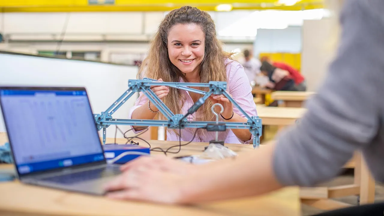 A woman smiles while assembling a metal structure at a table, with a laptop in the foreground and another person working nearby.