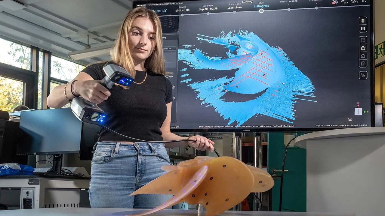 A person is standing up holding a hand held scanning device which is aimed at an item on a table, the results of the scan are on the tv screen behind them