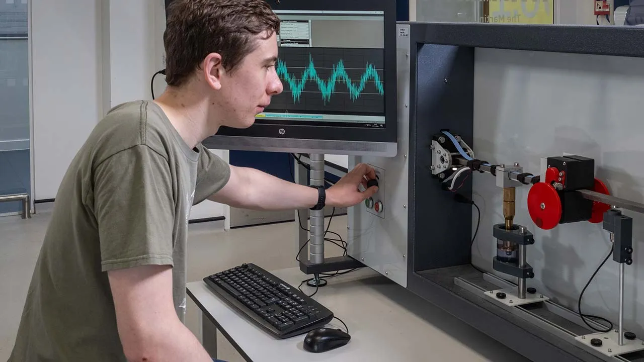 A student is sat at a desk and pressing a button to operate a mechanical device. There is a computer screen showing data in the background