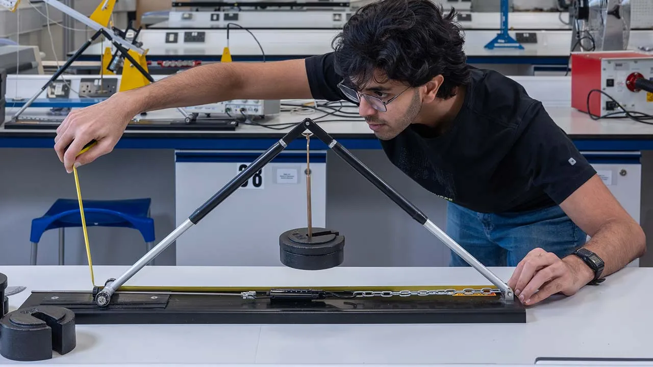 A student is working at a bench in a lab and measuring a device that is in front of the with a tape measure