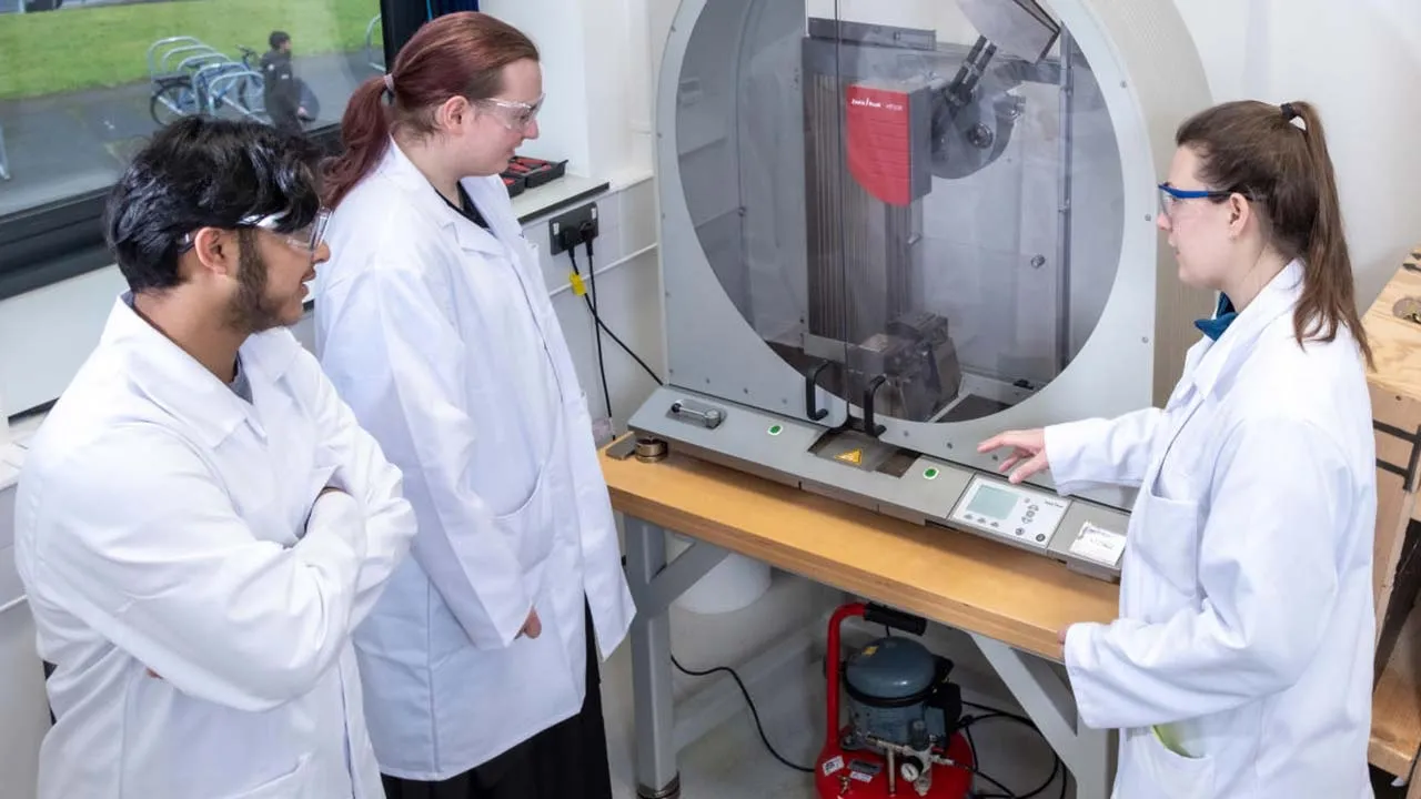 Three students in lab coats examining a complex machine in a laboratory