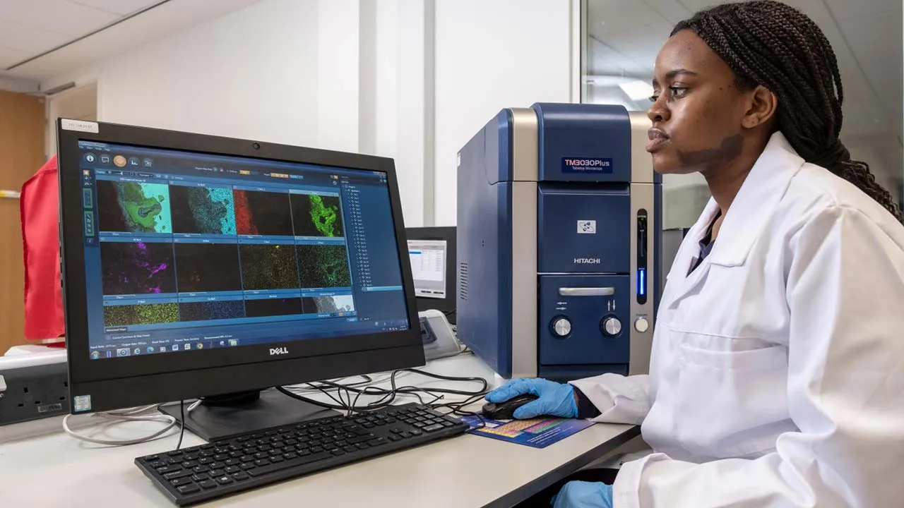 A student wearing a lab coat and gloves is using a computer and looking at data on the screen
