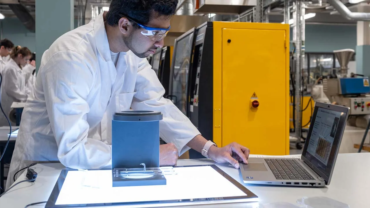A student wearing a lab coat and protective eye glasses has a magnifying lens in front of them and is using their laptop