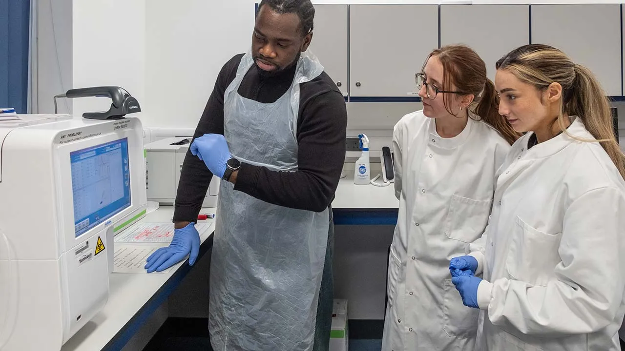 Three students in a lab wearing protective clothing are looking a computer screen on a machine