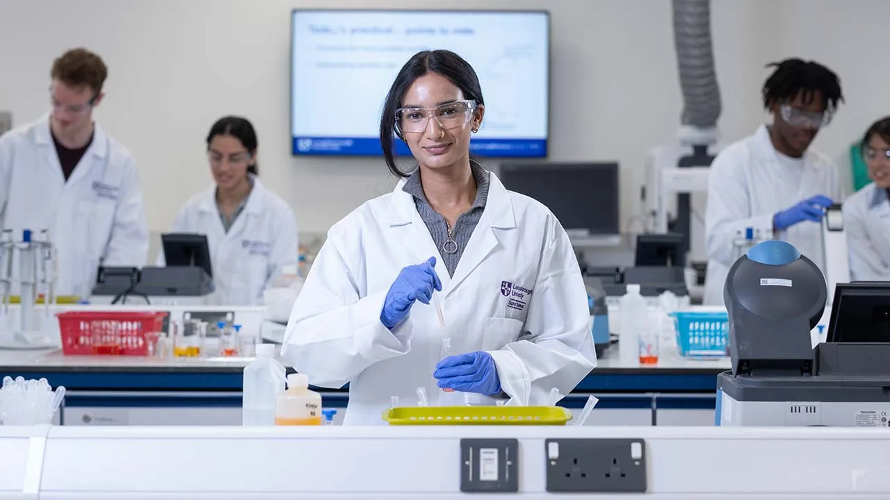 A chemistry student wearing a lab coat, protective glasses and gloves is carrying out an experiment in a lab. They are holding a test tube and there are other students working in the background