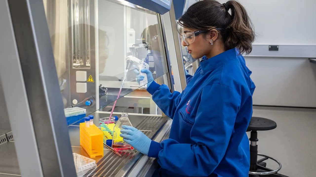 A chemistry student wearing a blue lab coat, protective glasses and gloves is mixing liquids behind a protective shield.