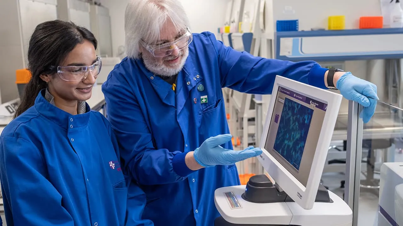 Two individuals in a chemistry lab wearing blue lab coats are looking at a computer screen