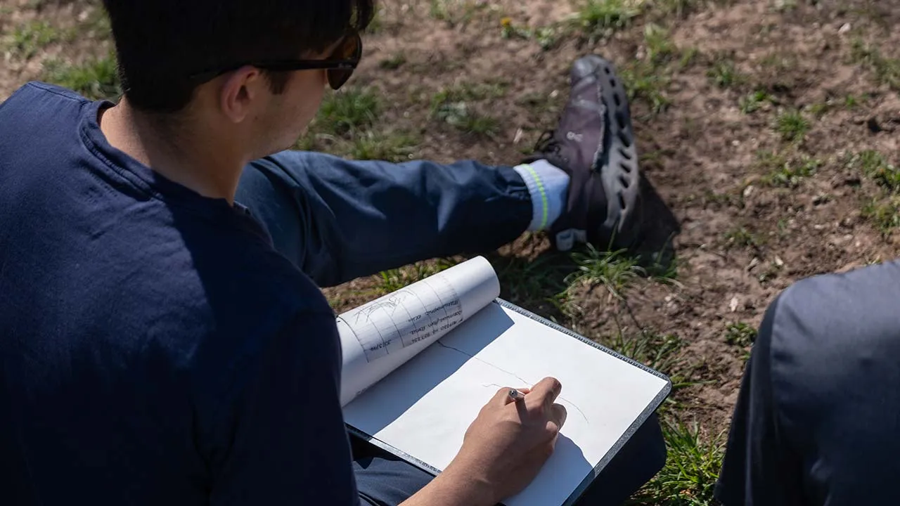 A geography student sat on the ground outside drawing with pen and paper