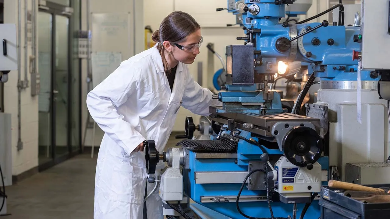 A student wearing a white lab coat is operating some machinery in a lab