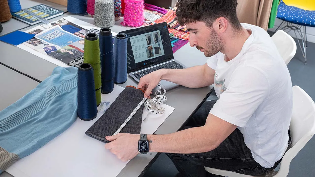 A design student working at a desk that has different yarns and a laptop on. They are using a measuring tape to measure some material