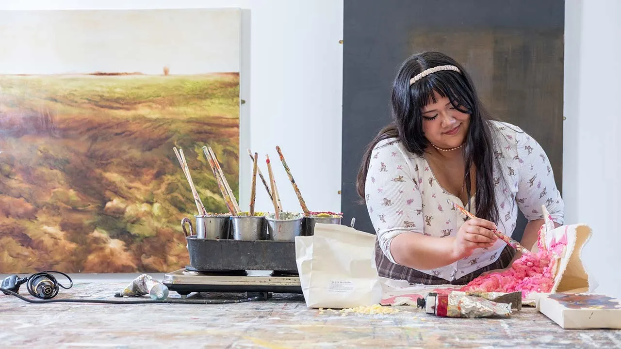 A woman is painting at a work bench. There are large painted canvases and paint supplies around her