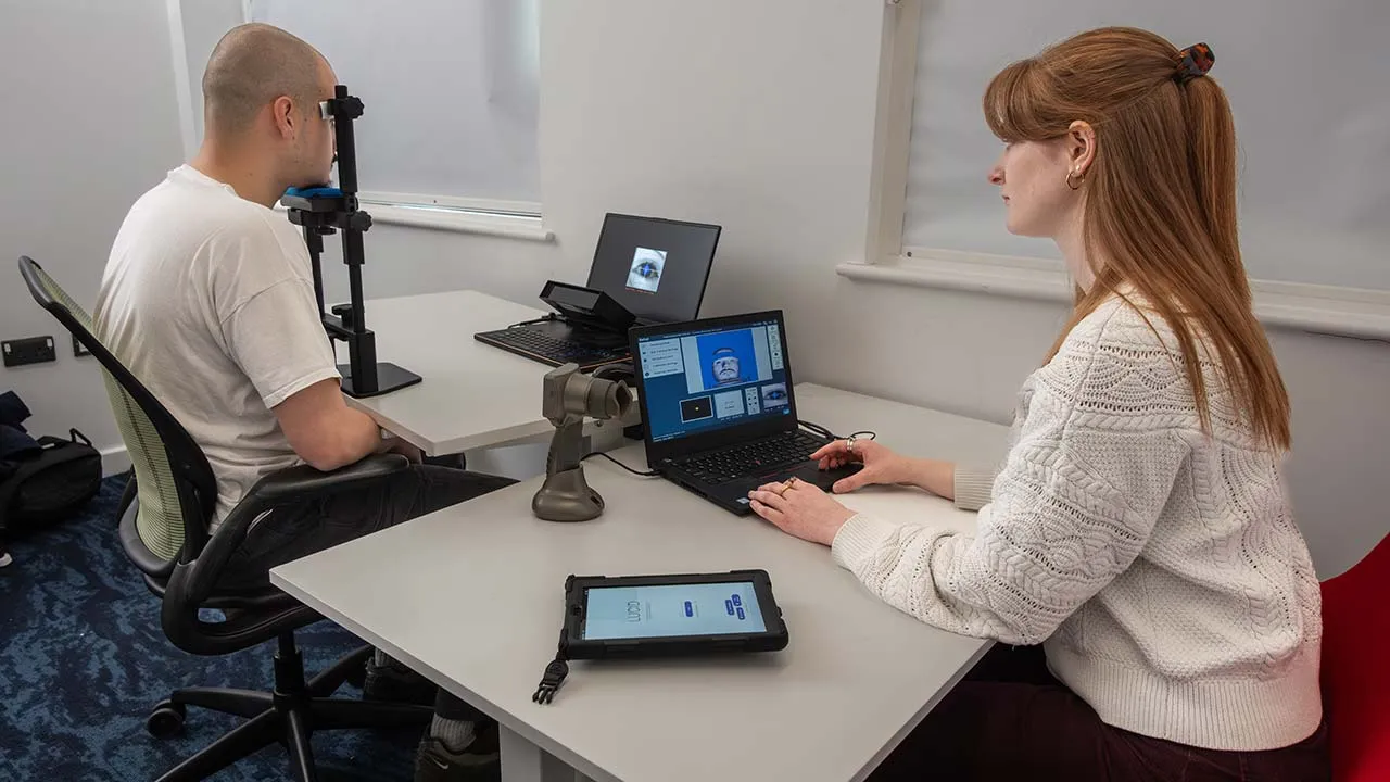 One person has their face on a head rest and is having a eye scan whilst another person is looking at the images on a laptop screen