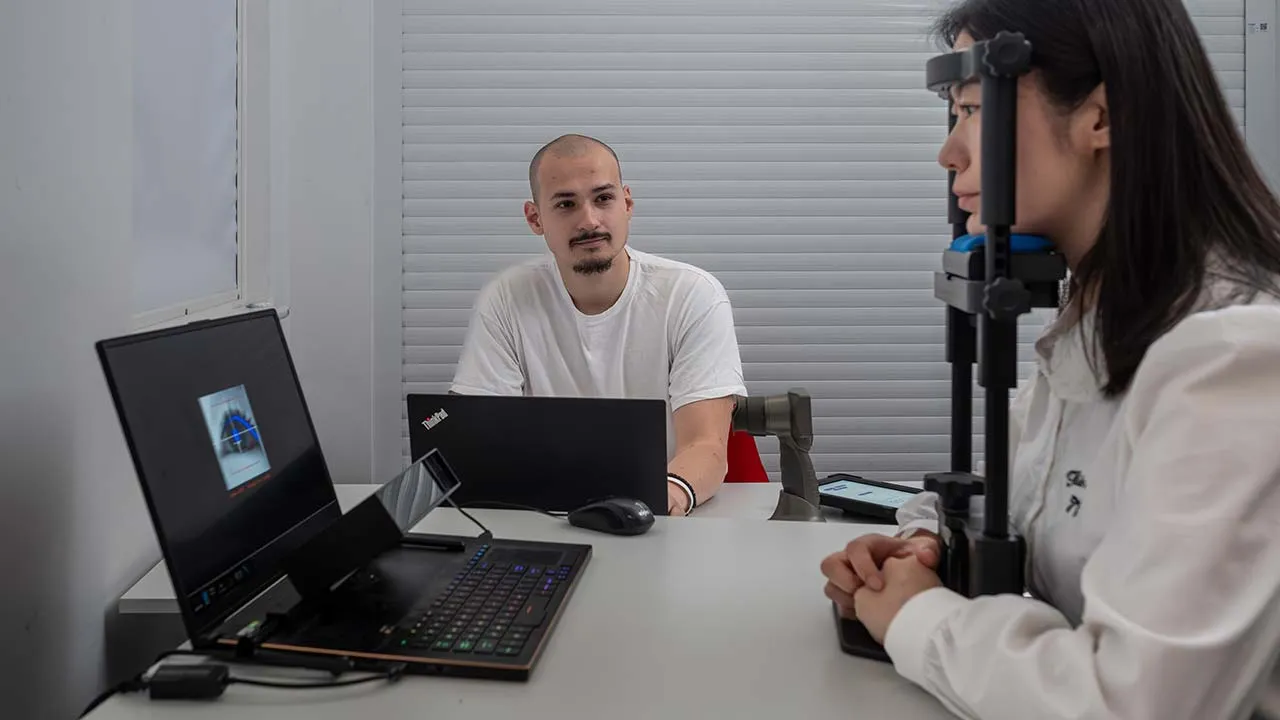 One person has their face on a head rest and is having a eye scan whilst another person has a  laptop open in front of them