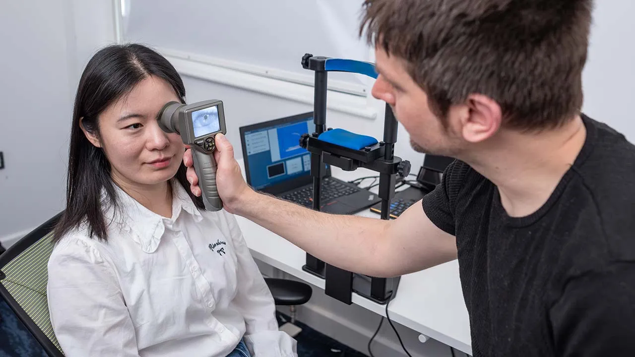 One person is holding an hand held device over the eye of another person who is sitting down