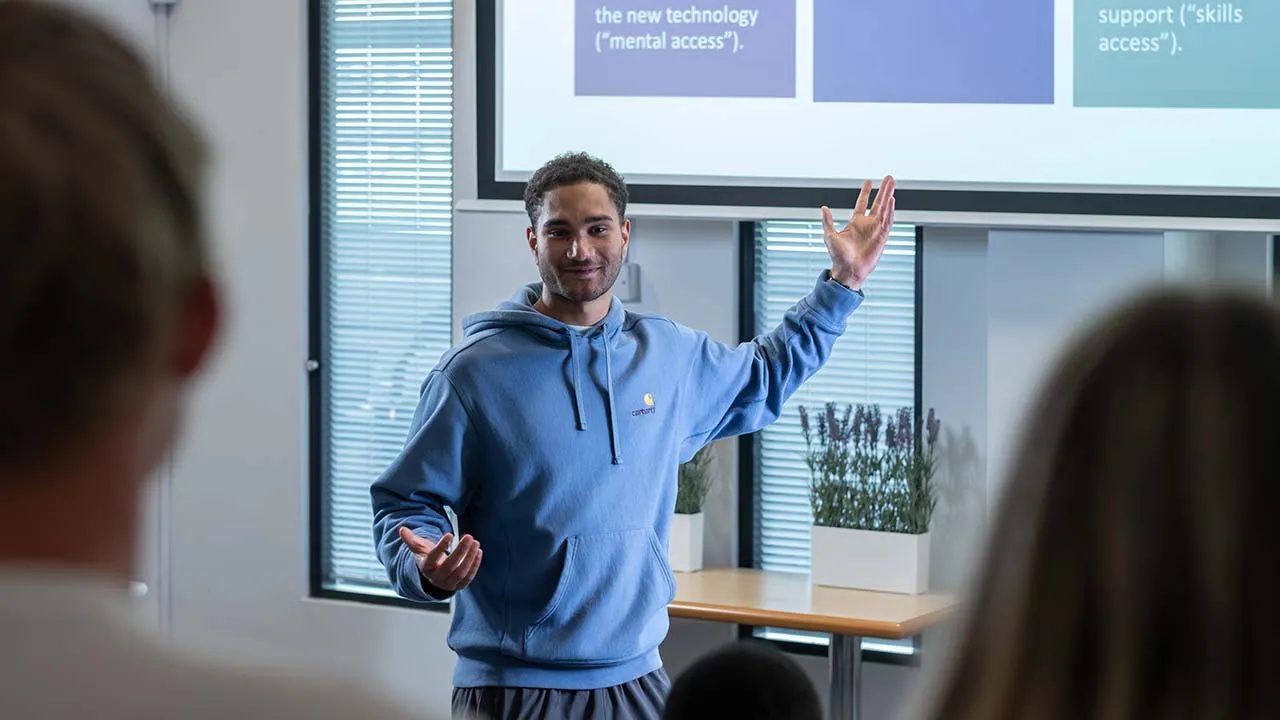 A man stood presenting to students who are sat facing him. There is a projector screen in the background that the presenter is pointing to