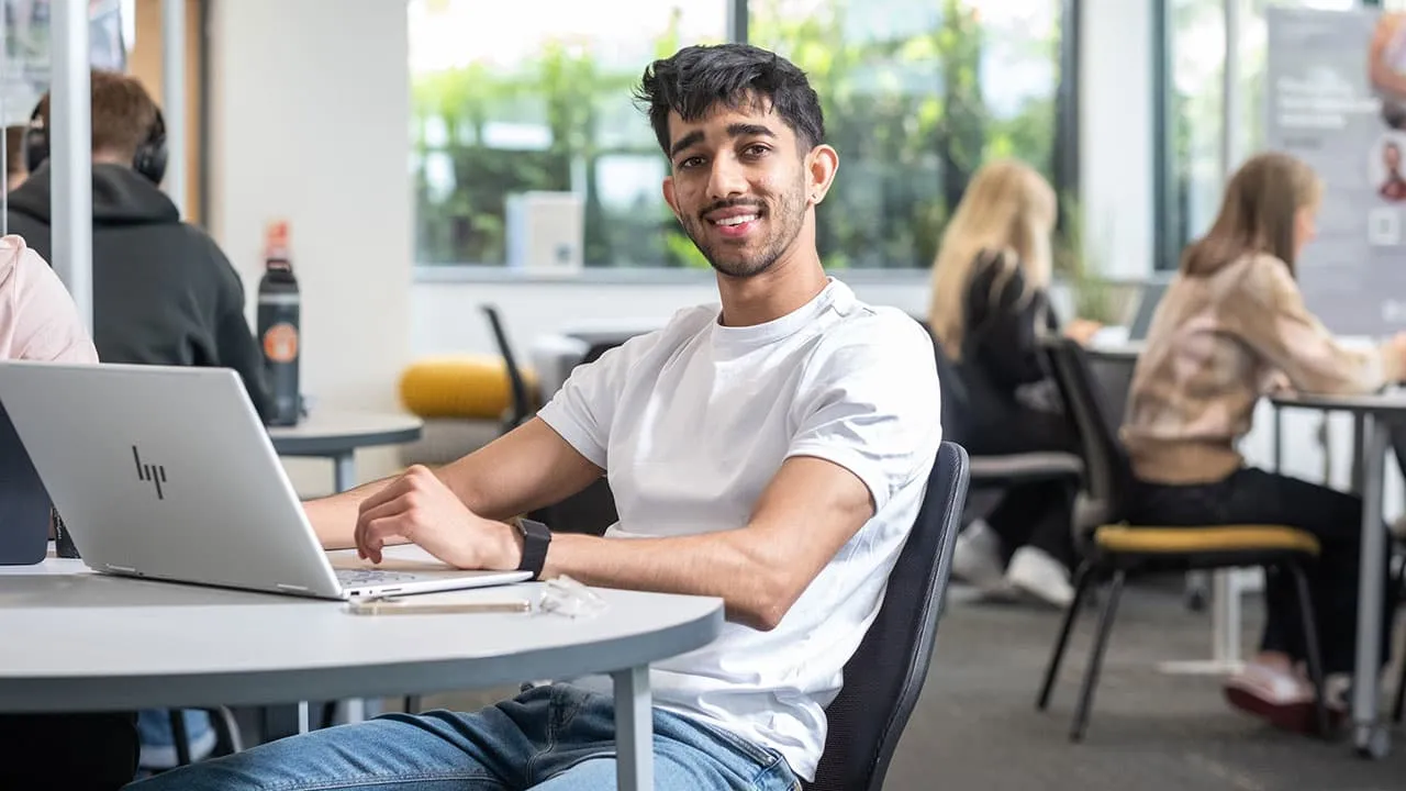 A student seated at a table, using their laptop. There are other students working in the background