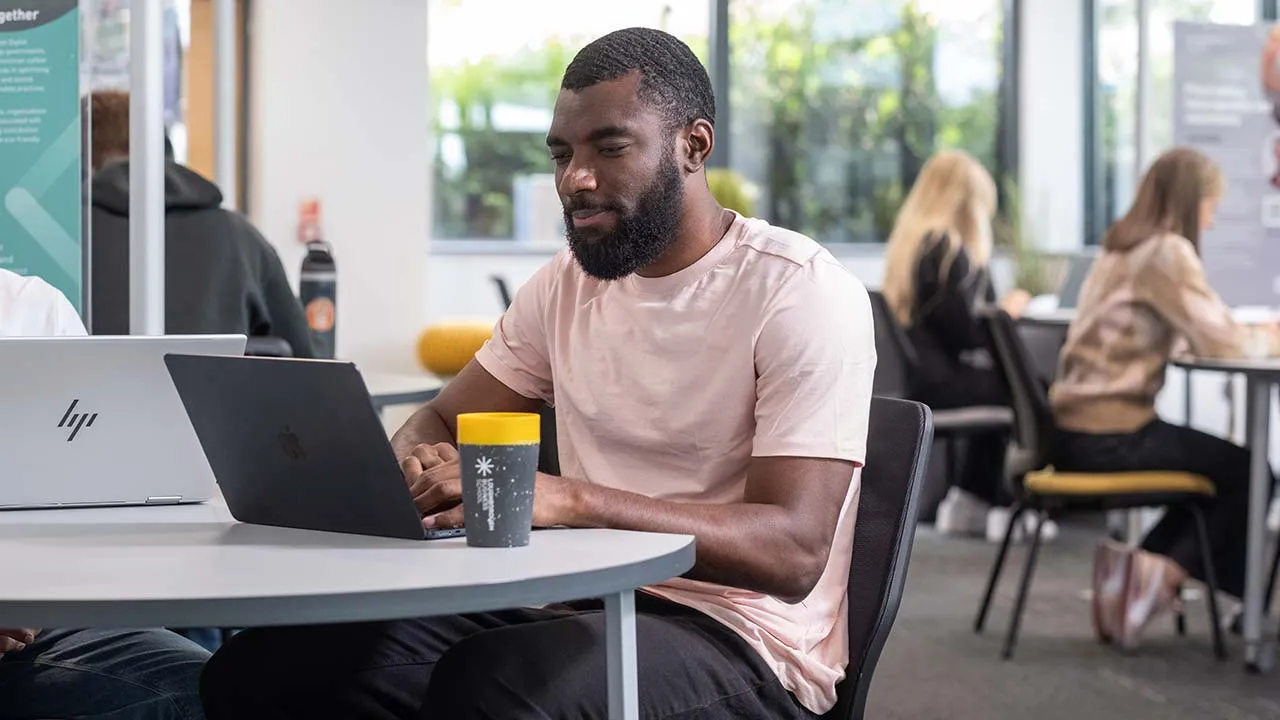 A man sat working on a laptop in a shared study space. Other students are sat working in the background