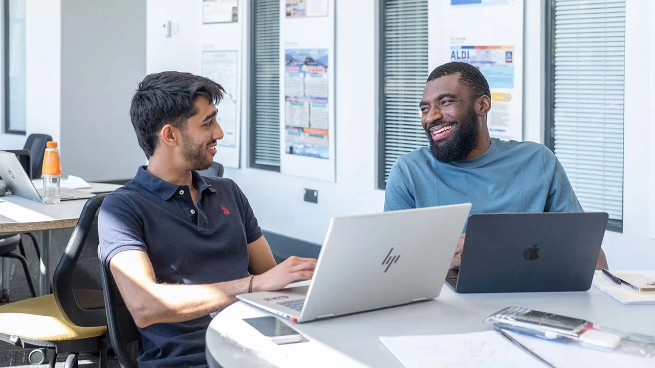 Two students sat at a desk talking to each other and smiling. They have laptops open on the desk