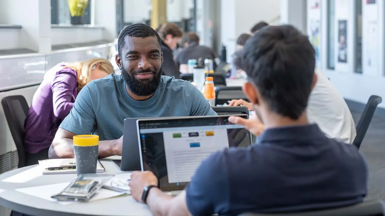 Two students sitting at a table working on a laptop and talking to each other