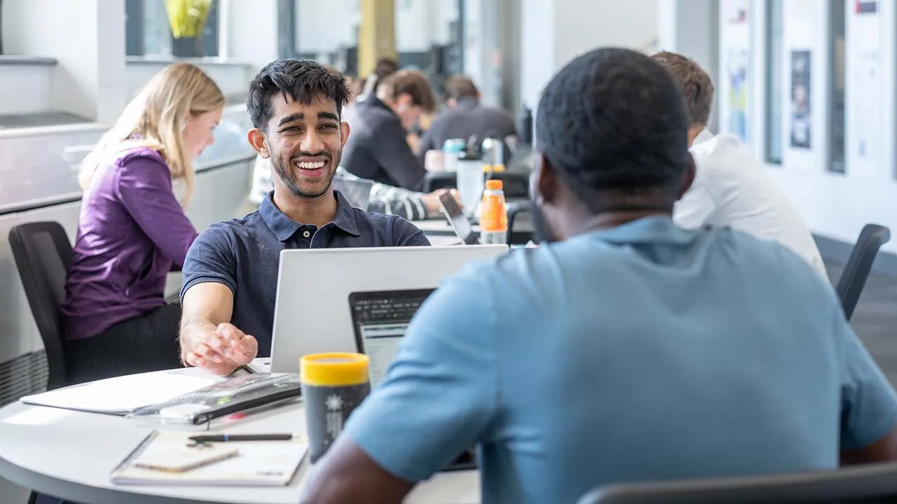 Two students sitting a table working in a shared study space. They have a laptop, paper and pens in front of them and there are others working in the background