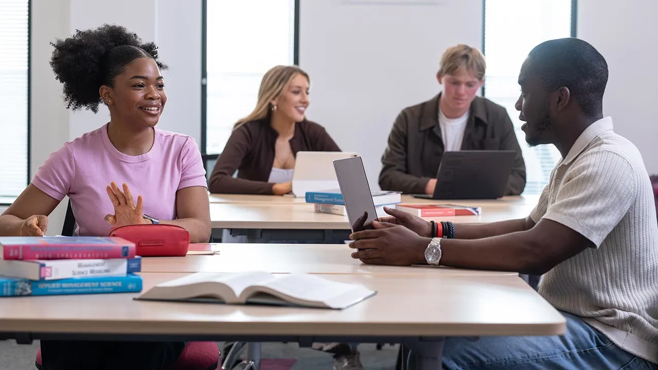 Four students sat at tables in a shared working space. There are laptops and books in front of them