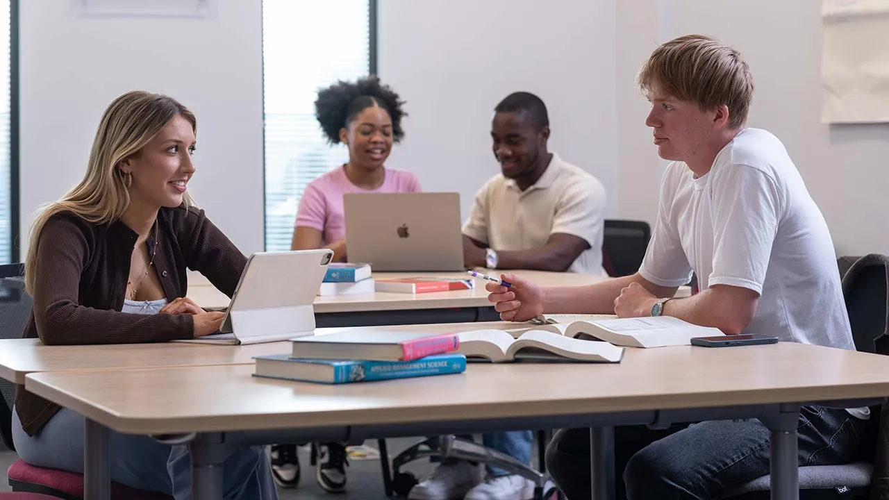 A group of students working in a shared study space with laptops, books and pens on the tables in front of them