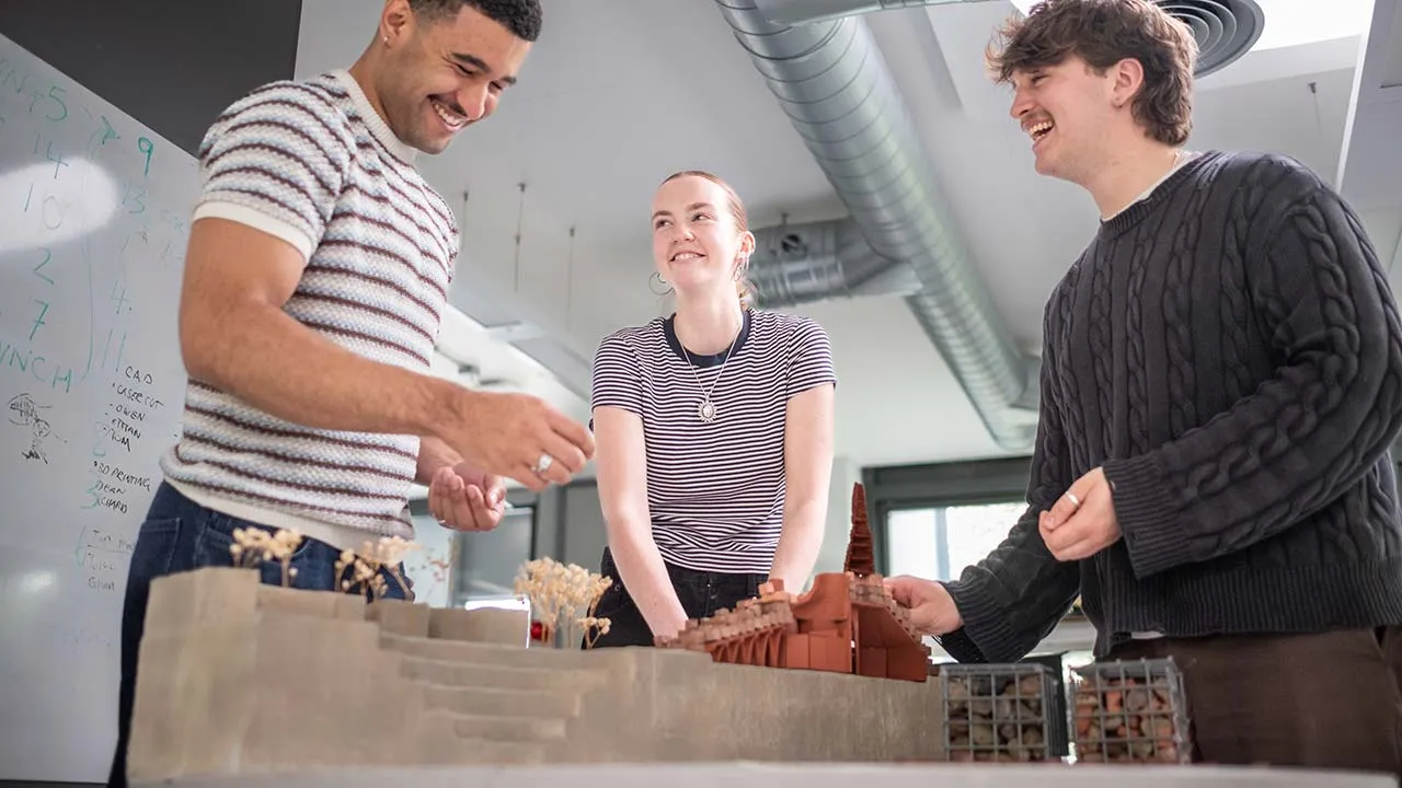 Three students working on a building model together and smiling at one another
