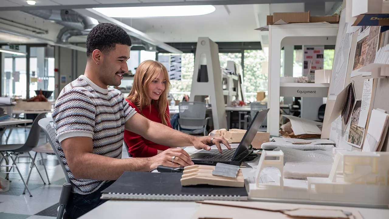 Two students collaborate on a laptop in a bright workspace with technical drawings on the desk in front of them