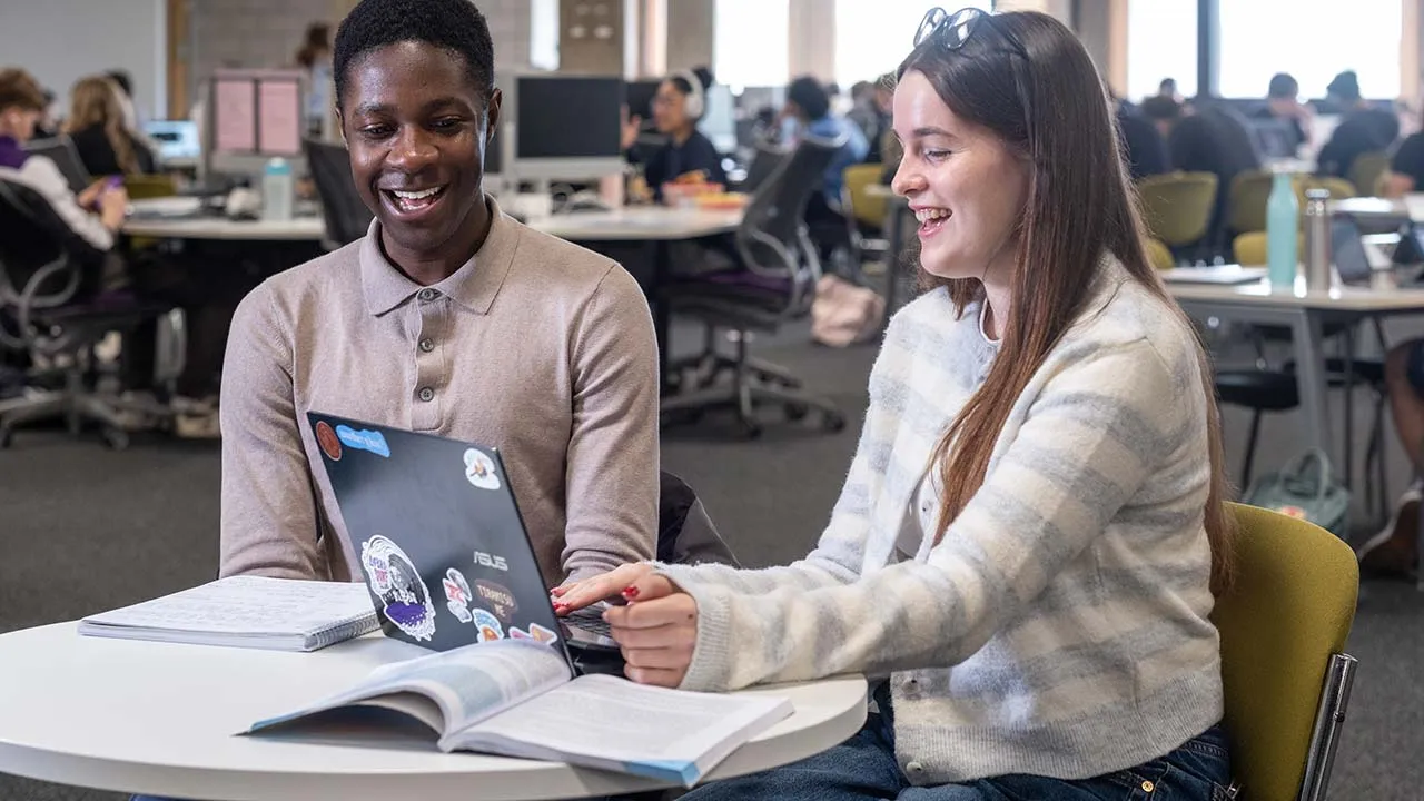 Two students are sat at a table working in a shared study area. There is a laptop and books in front of them