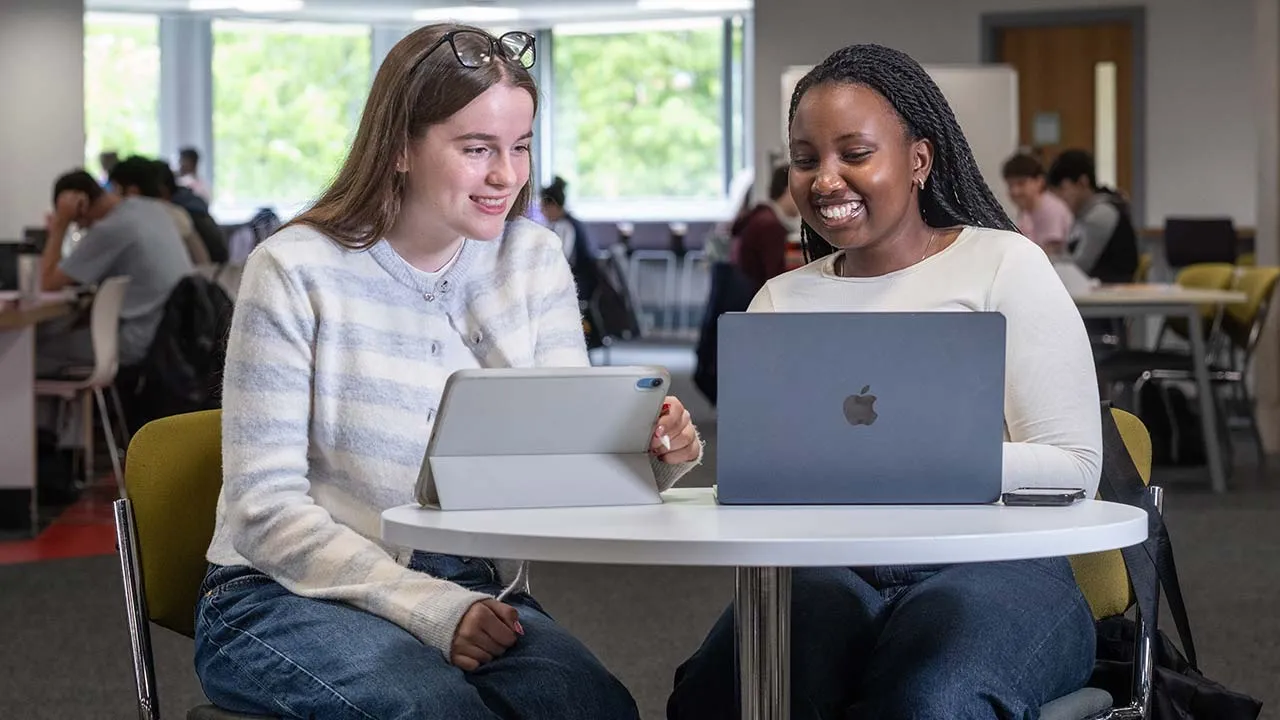 Two women sat at a table with iPads in front of them. There are other students sat working in the background