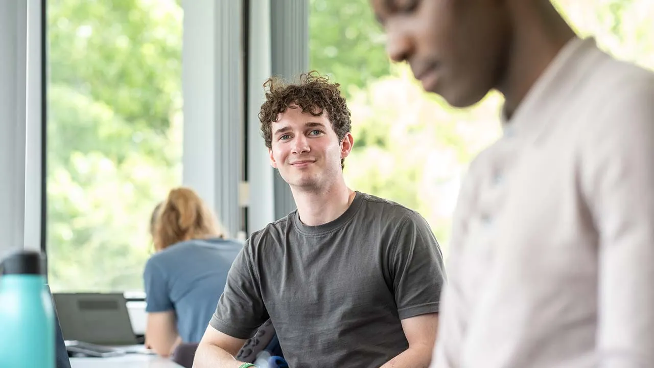 Three students are sat at desks working. Two are looking down and reading, the other is looking towards the camera