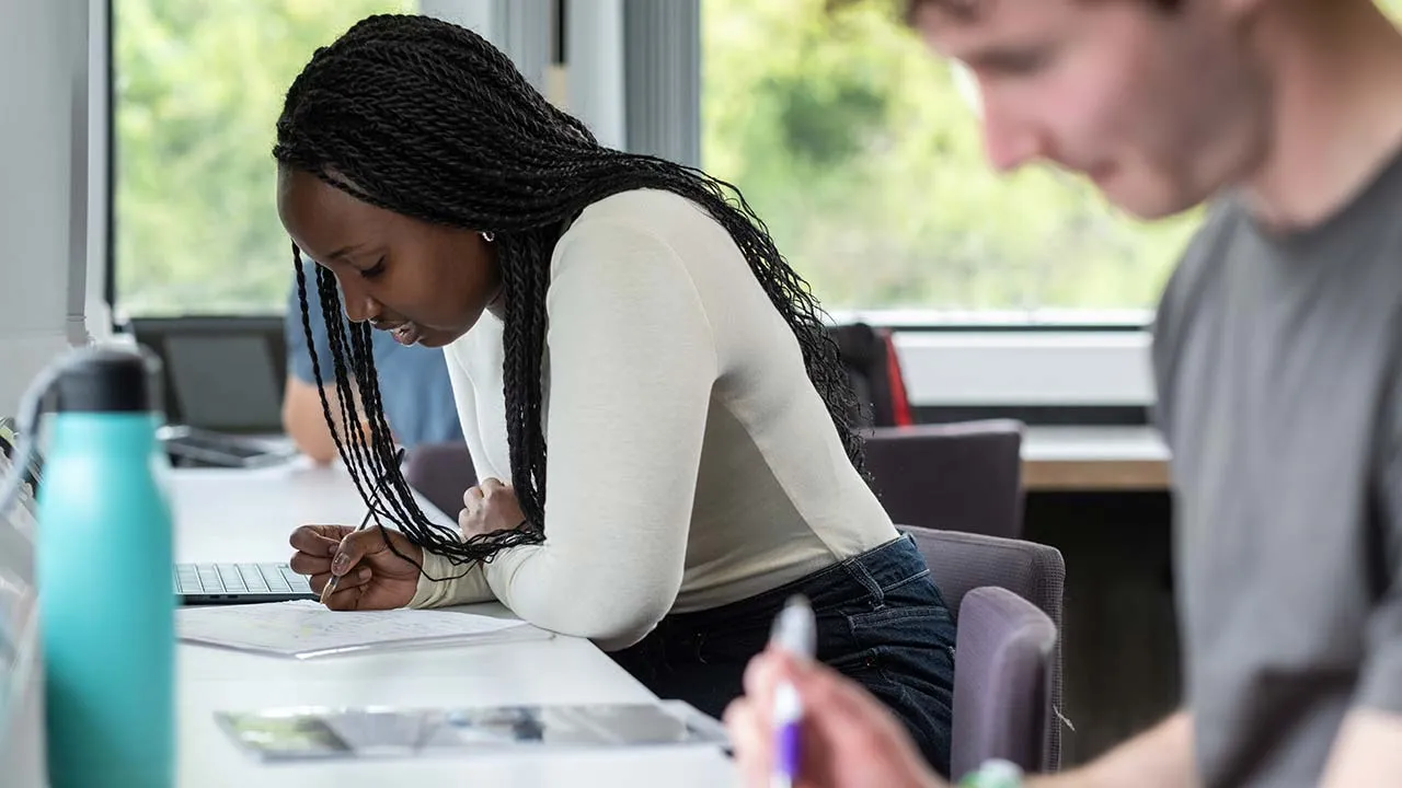 Two students are sat next to each other writing with pen and paper