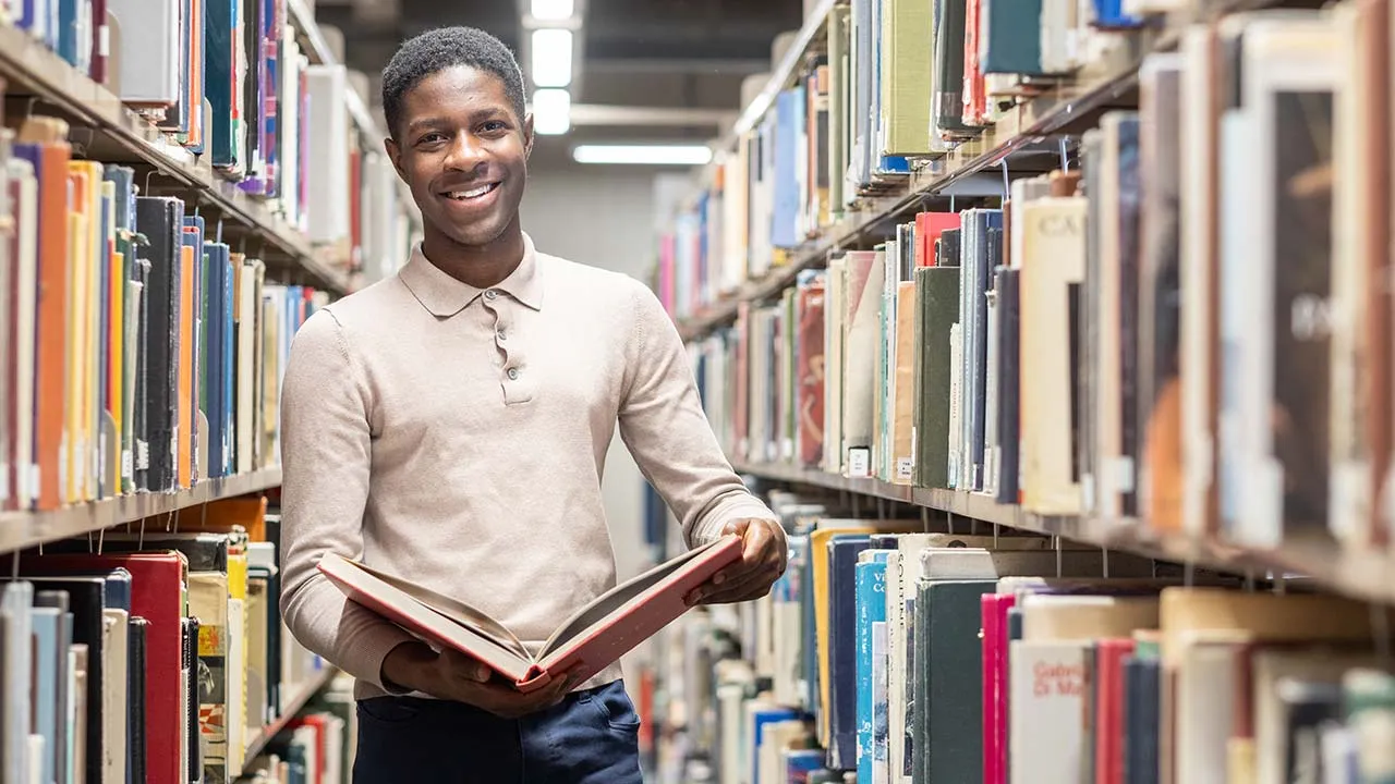 A man stood in the library holding an open book. There are rows of books on shelves around him