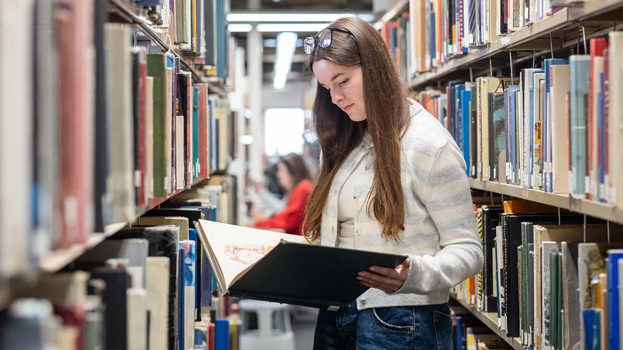 A student stood reading an open book in the library. There are shelves of books in the background