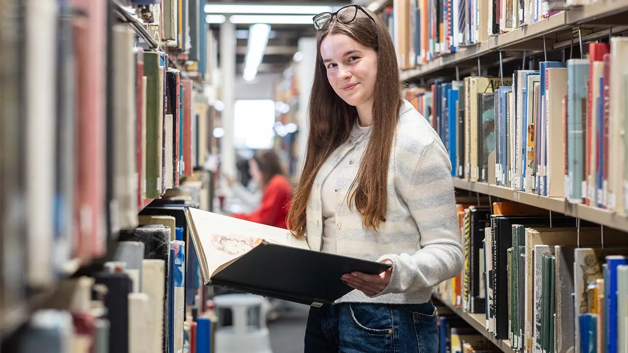 A female student holding a book whilst standing in an aisle in the library. There are rows of books on the shelves around her.
