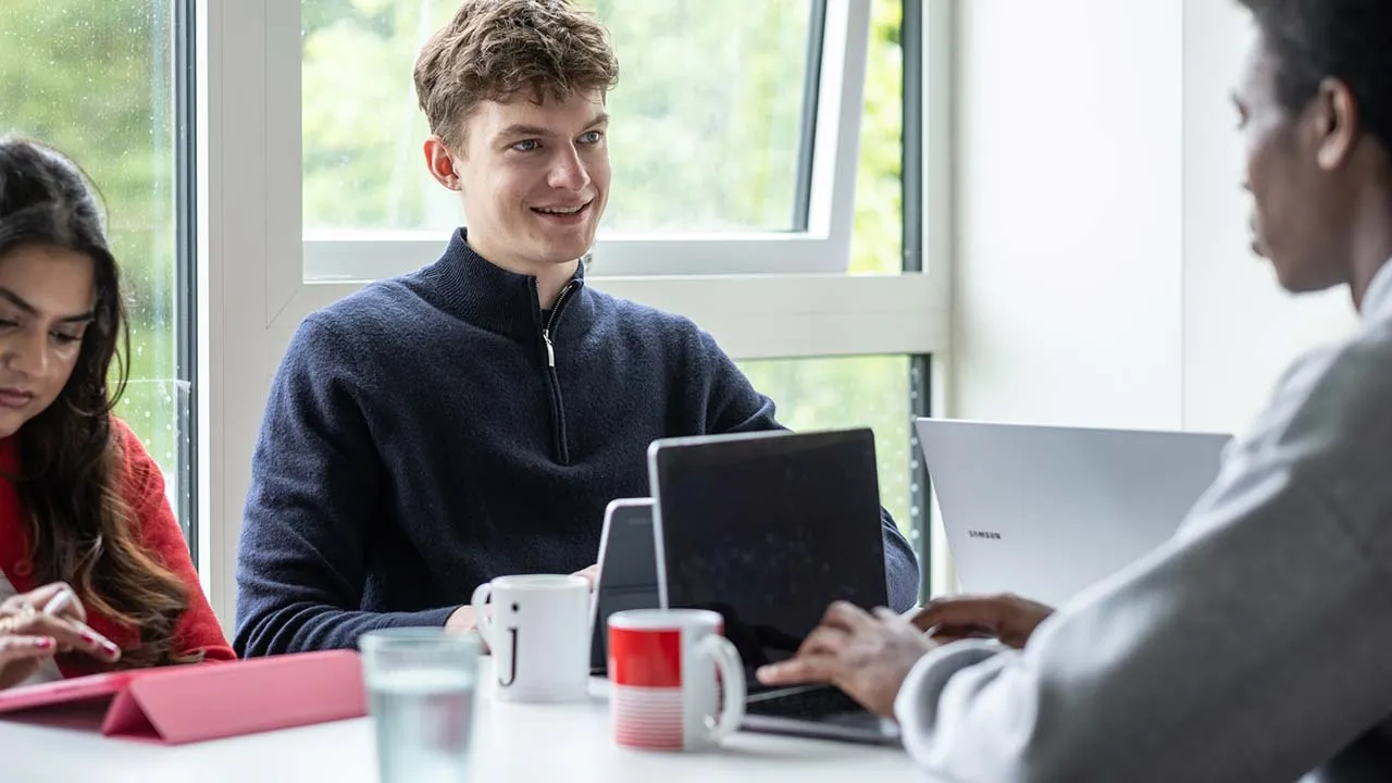 Three students are sat working at a table. There are laptops, iPads and drinks on the table