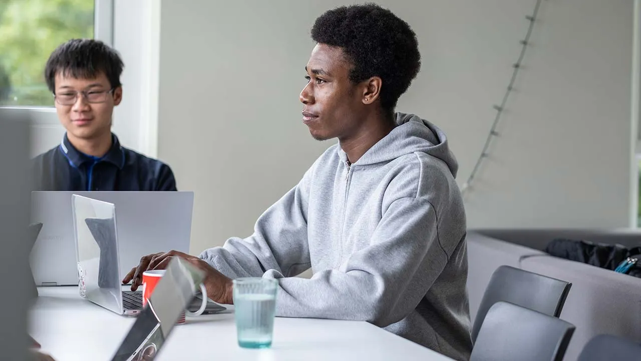 A student is sat working on a laptop. There is a glass of water next to them