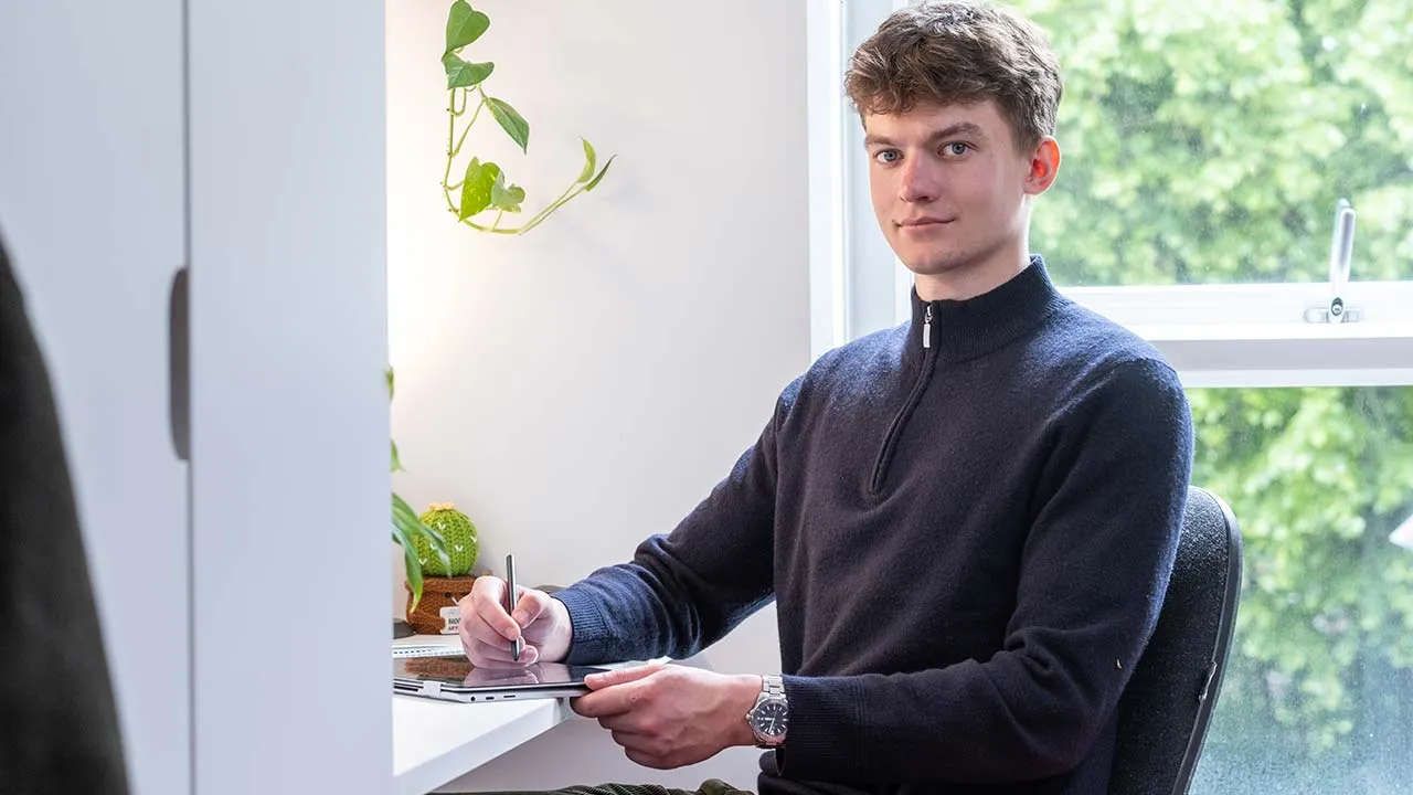 A student sat at a desk in their student accommodation writing on a tablet device