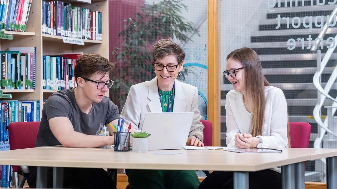Three people sat at a table talking, one with a laptop in front of them. There are maths and statistics books in the background