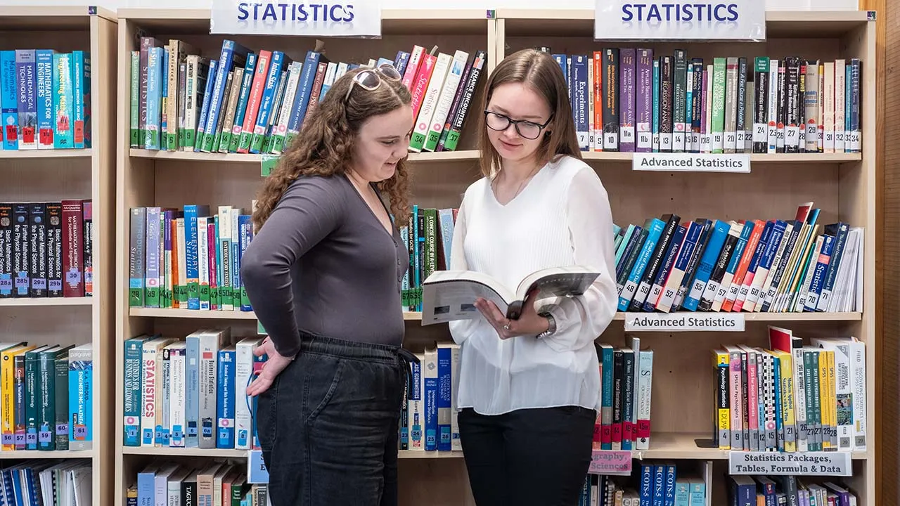Two people stood looking at a book with shelves filled with statistics books behind them