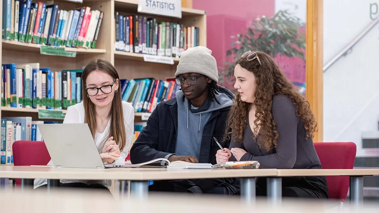 Three students sat at a desk working on a laptop. There are maths an statistics books in the background