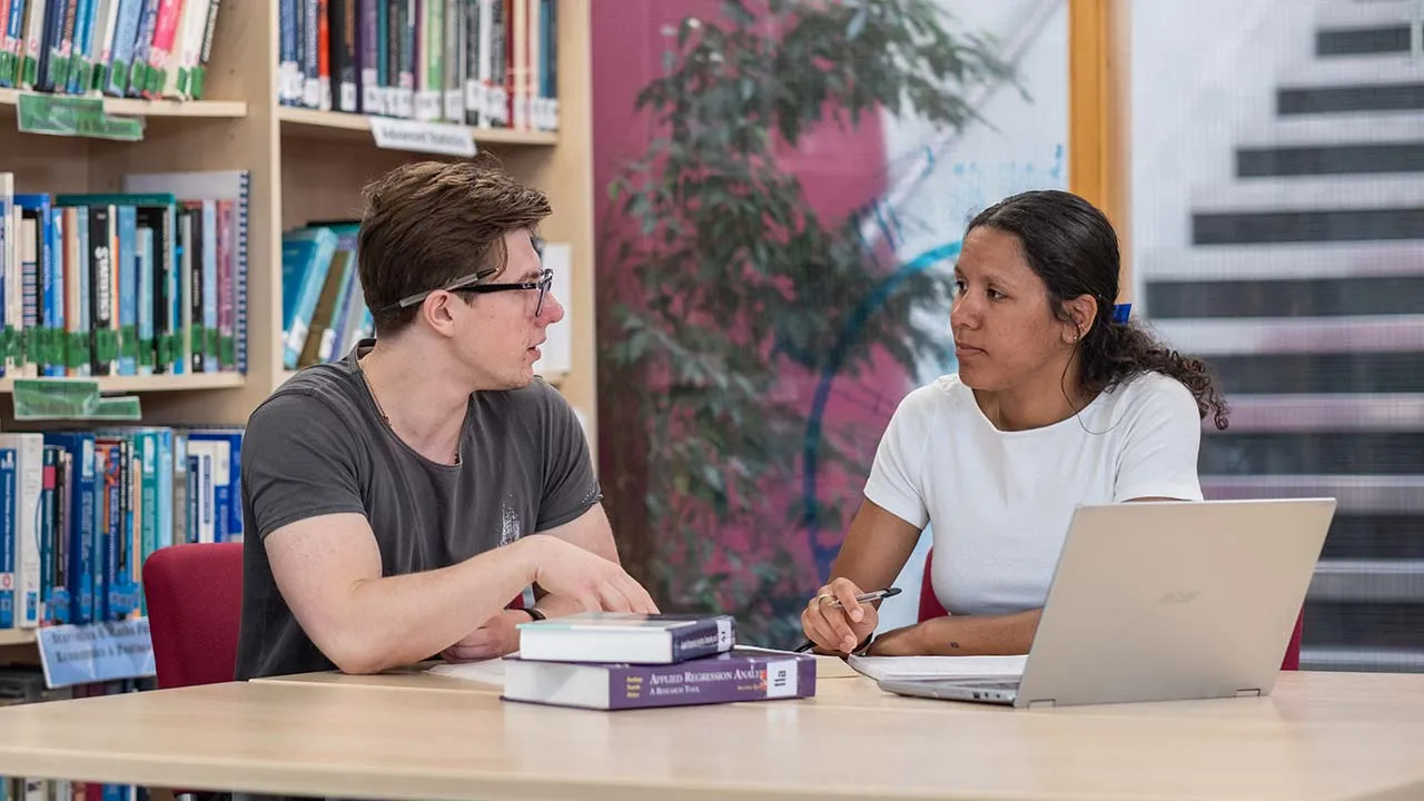 Two people sat talking. There are books and a laptop on the table in front of them and maths books on the shelves behind them