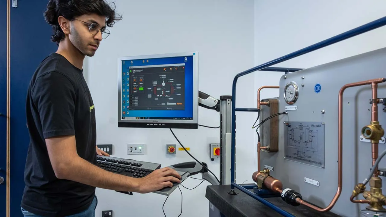Person operating a computer-controlled engineering test rig fitted with copper piping, pressure gauges, and refrigeration system components.