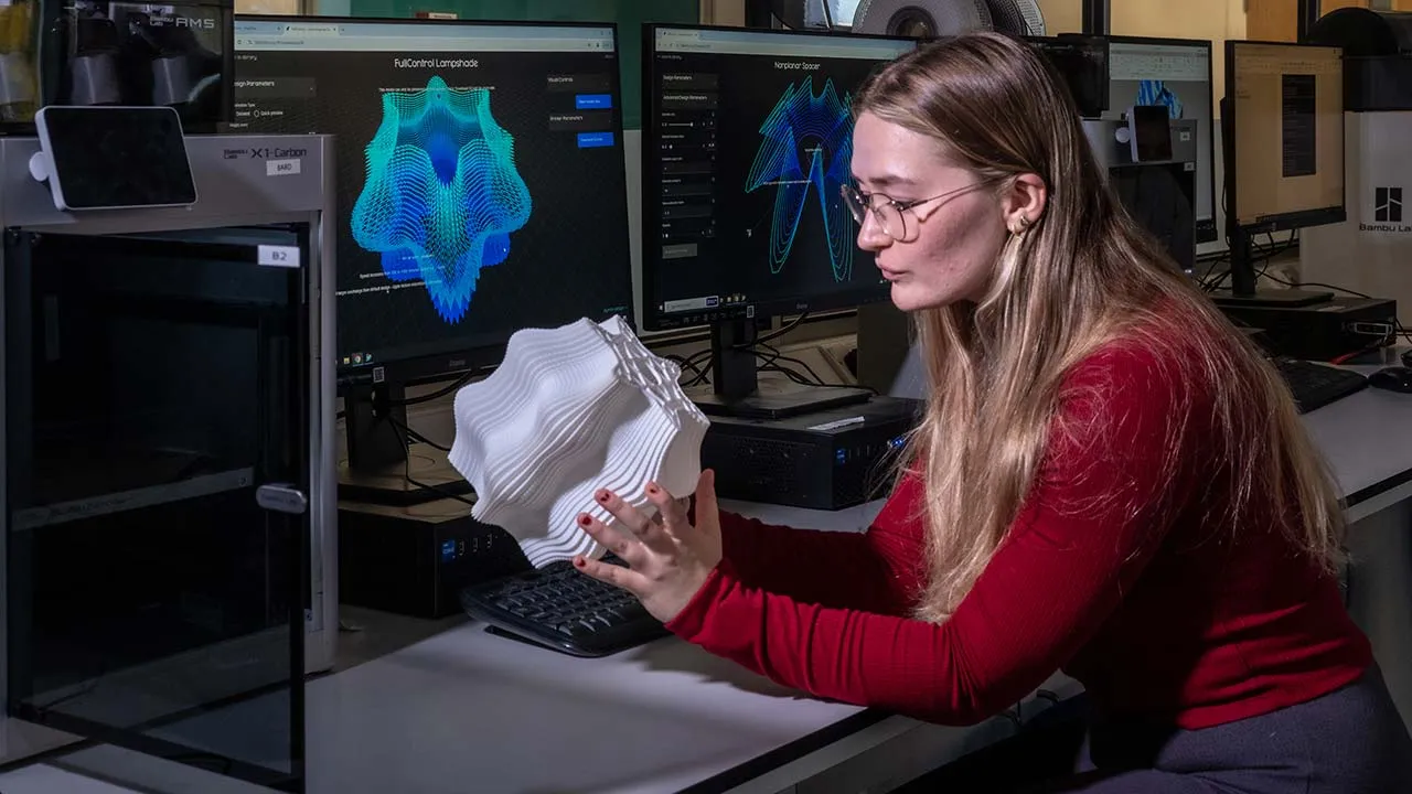 Person seated at a workstation in a digital fabrication lab, holding a white 3D-printed sculptural model