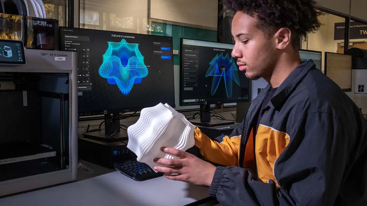 Person seated at a workstation in a digital fabrication lab, holding a white 3D-printed sculptural model