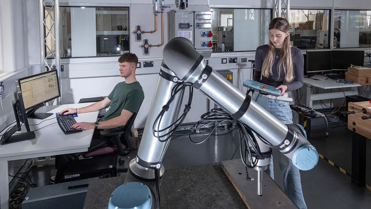 Modern robotics laboratory featuring a collaborative robotic arm in the foreground. One person operates software at a computer workstation while another uses a handheld control device beside the robot.