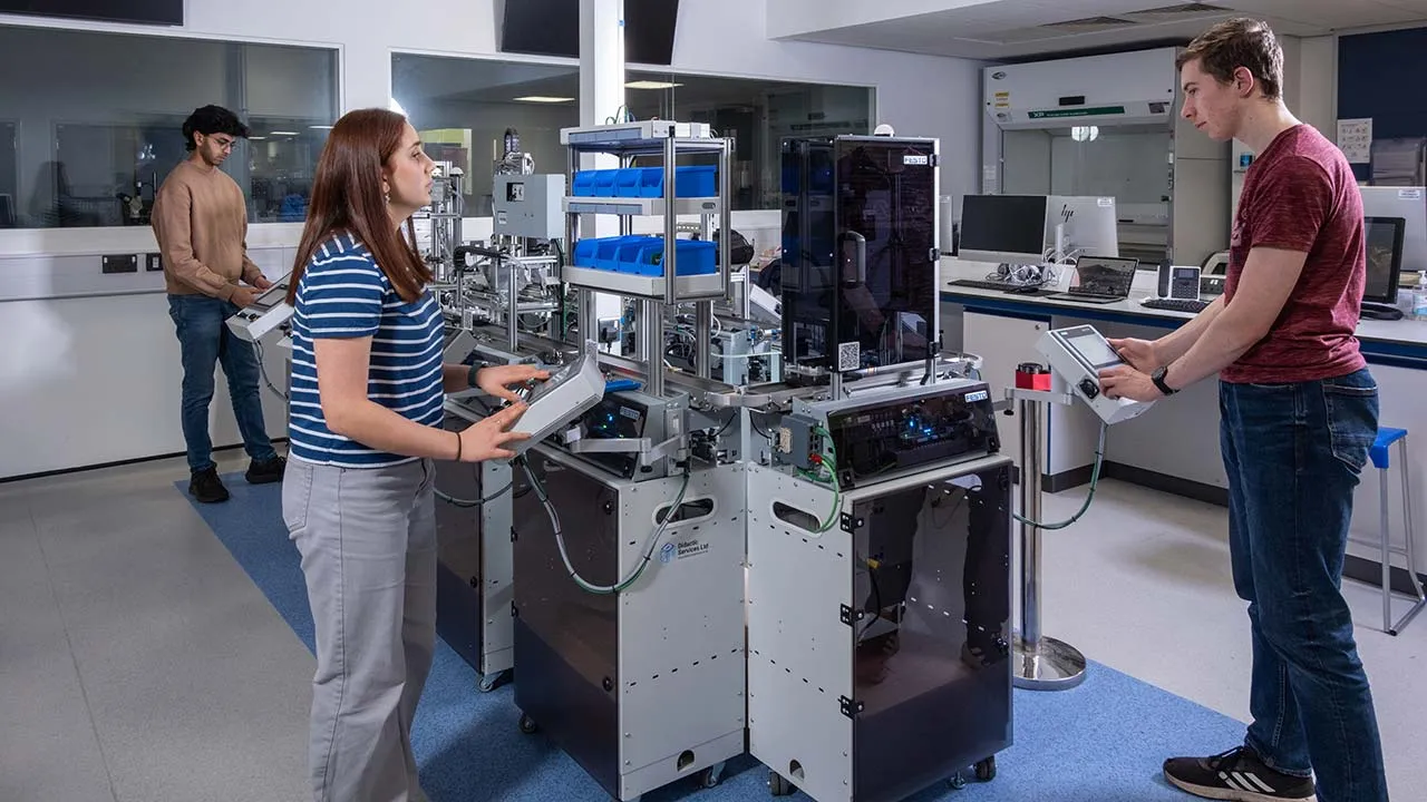 Three people operate an automated manufacturing and robotics system in a laboratory environment.