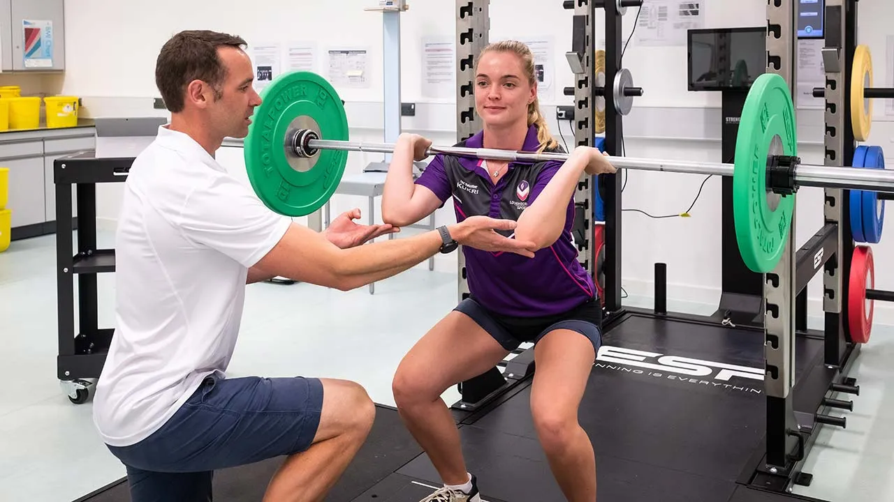 One person is lifting weights whilst another person is standing close by to spot them